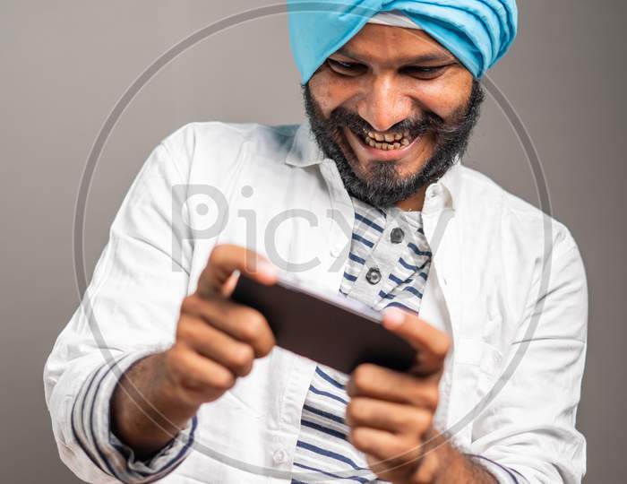 Young Smiling Sikh Man Busy Playing Video Game On Mobile Phone - Concept Gaming Addiction, Using Modern Technology On Studio Background.