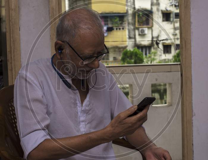 An Old Man Busy Listening Music From His Smart Phone And Blue-Tooth Headphone. Concept Of Technology For Everyone. Selective Focus.
