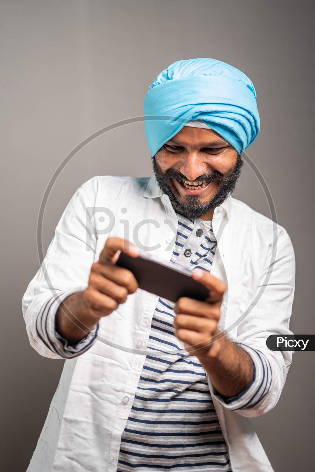 Young Smiling Sikh Man Busy Playing Video Game On Mobile Phone - Concept Gaming Addiction, Using Modern Technology On Studio Background.