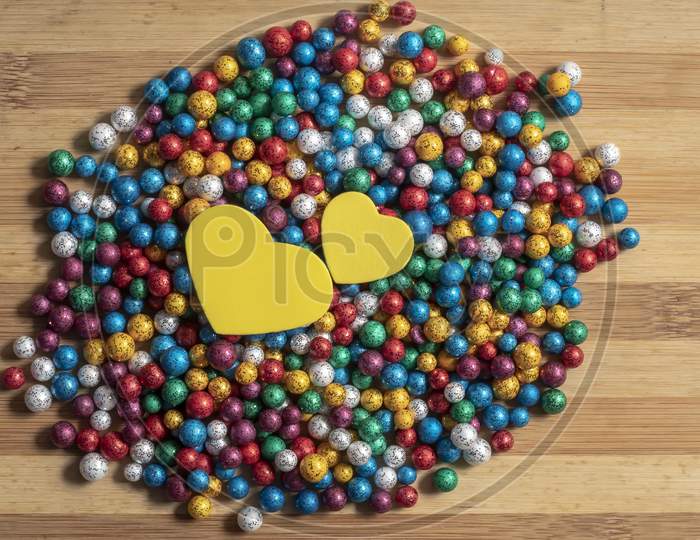 Valentine'S Day Background. Multicolor Small Size Thermocol Decorative Balls With Two Yellow Hearts On Top Of A Wooden Table