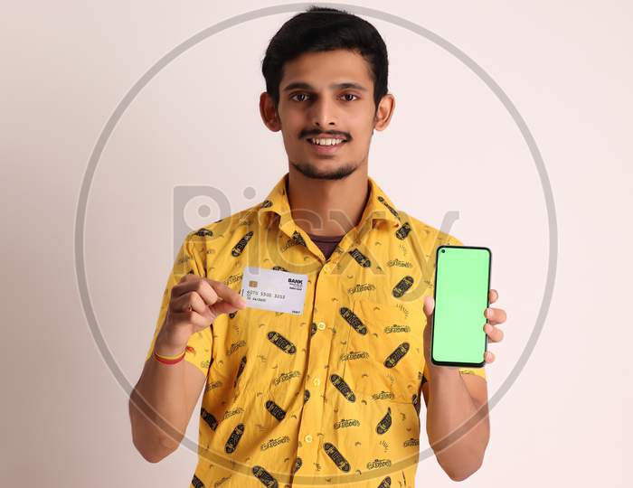 Young Smiling Bearded Attractive Indian Boy Wear Professional Yellow Basic Shirt Holding Mobile Cell Phone And Credit And Debit Card of Bank for Shopping Online On Isolated Light Brown Background