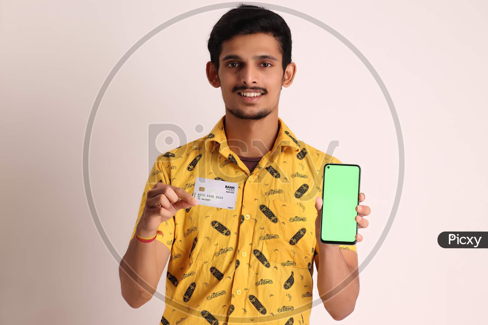 Young Smiling Bearded Attractive Indian Boy Wear Professional Yellow Basic Shirt Holding Mobile Cell Phone And Credit And Debit Card of Bank for Shopping Online On Isolated Light Brown Background