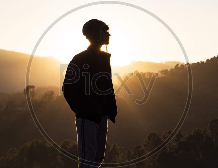 Silhouette Of A Young Boy Standing In The Fields And The Beautiful Mountains In The Background Captured During Sunset.