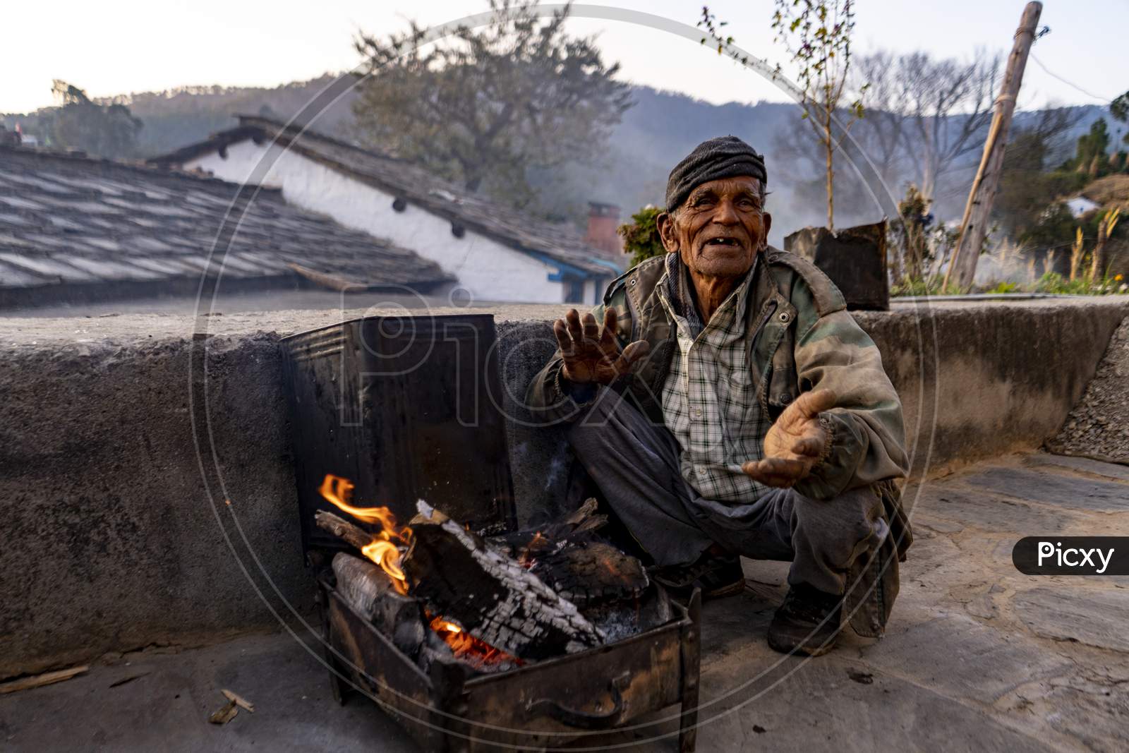 Almora, Uttarakhand - Jaunary 2 2022- An Old Man Sitting In Rugged Clothes, Soaking Warmth From Firewood. An Village Old Man Smiling Into The Camera.