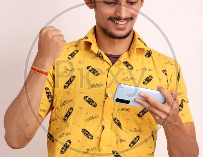 unshaven attractive Indian guy with wearing yellow shirt and Playing games on android smartphone on and gesturing emotion sign with face isolated on white background studio portrait seating on chair