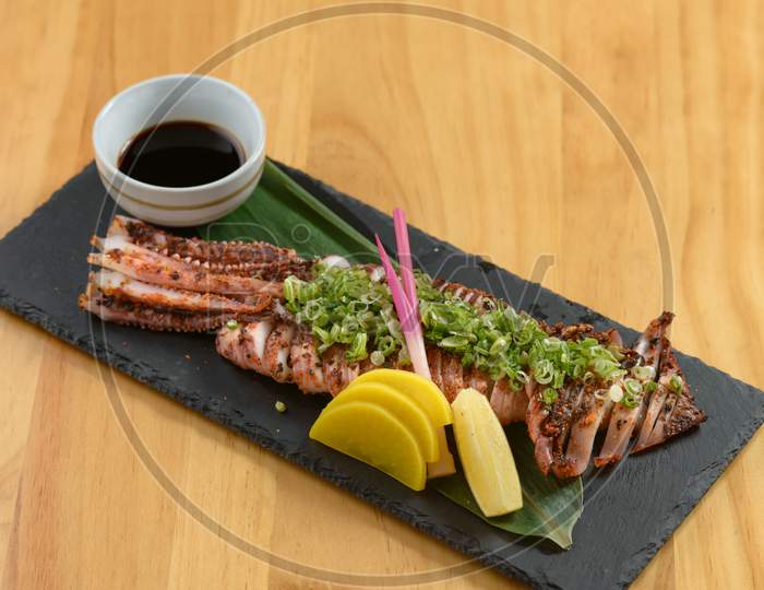 Japanese Food Grilled Surume Ika With Lemon And Soy Sauce In A Black Dish Isolated On Wooden Table Top View